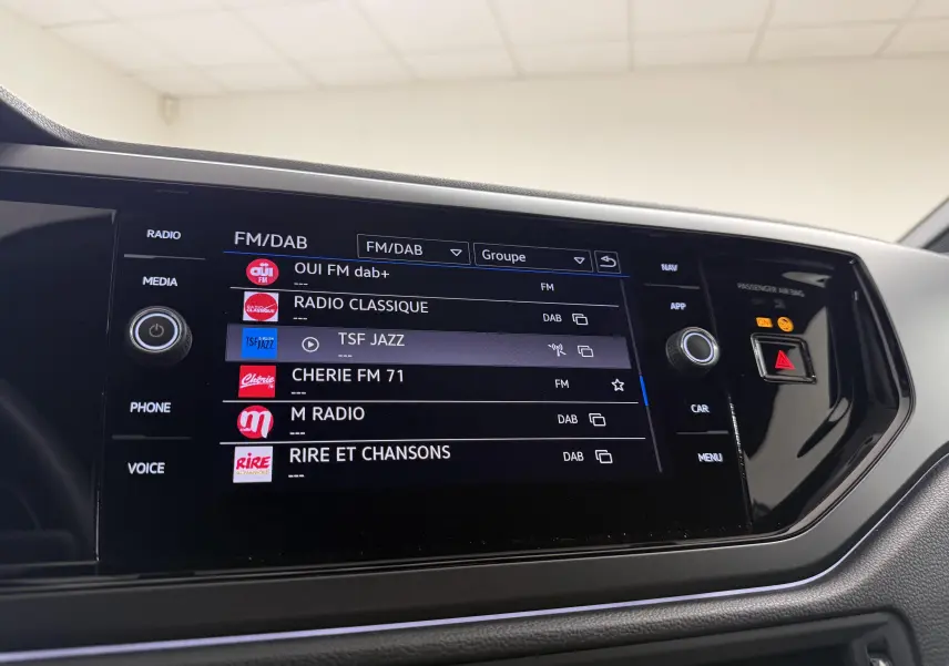 Écran tactile central du Volkswagen Taigo 2025 affichant les stations radio FM/DAB, avec commandes autour et finition noire brillante.