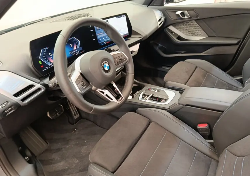 Vue intérieure côté conducteur de la BMW Série 1 120 DKG7 M Sport 2025, montrant volant cuir, écran tactile et sièges gris foncé.