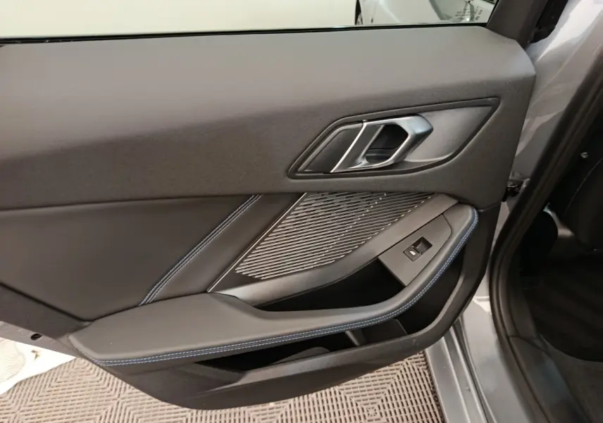 Gros plan sur la porte intérieure côté gauche gris clair de la BMW Série 1 120 DKG7 M Sport avec surpiqûres bleues et poignée métallique.