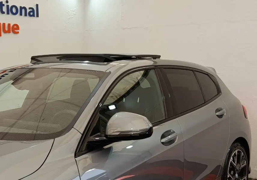 Vue de profil droit avant d'une BMW Série 1 120 DKG7 M Sport gris clair avec toit ouvrant et jantes sport noires.
