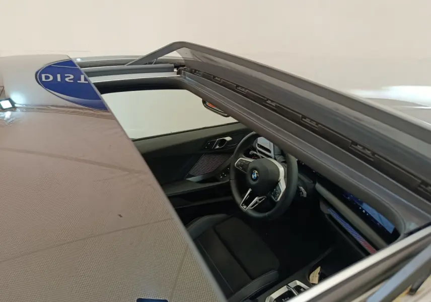 Vue intérieure par le toit ouvrant d'une BMW Série 1 120 DKG7 M Sport gris clair, volant multifonctions et tableau de bord numérique.