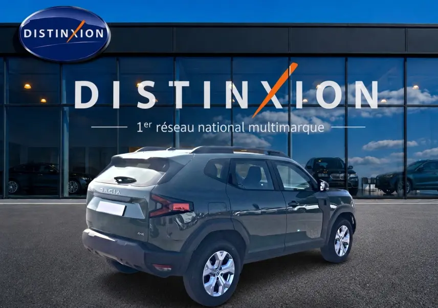 Vue 3/4 arrière droite d'un Dacia Duster 2026 kaki lichen avec barres de toit et jantes alu devant un showroom Distinxion.