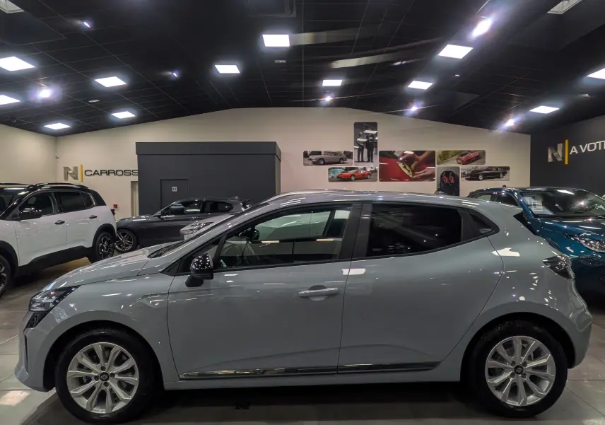 Profil côté gauche d'une Renault Clio V 2025 gris Rafale en intérieur, avec jantes alliage et rétroviseurs noirs.