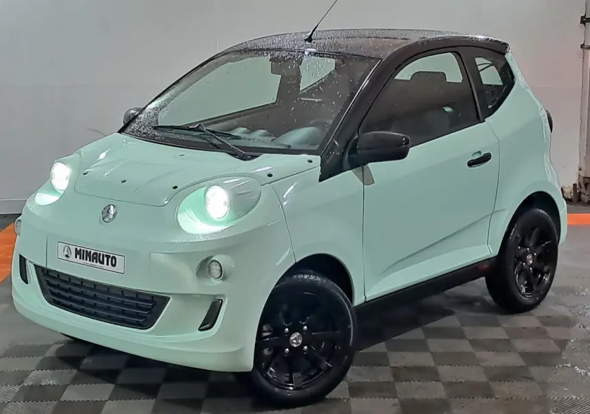 Vue 3/4 avant d'une Aixam Minauto E chic 2026 vert clair avec toit noir et jantes noires, phare avant allumé.