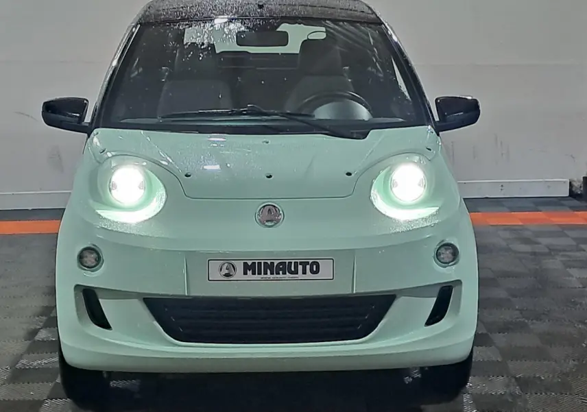 Vue frontale d'une AIXAM MINAUTO E chic 2026 verte électrique avec phares allumés et rétroviseurs caméra côté conducteur.