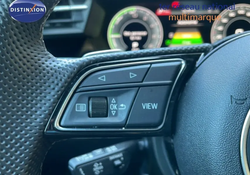 Gros plan sur les commandes à gauche du volant perforé noir de l'Audi A3 Sportback 40 TFSIe 2025 avec tableau de bord numérique flou.