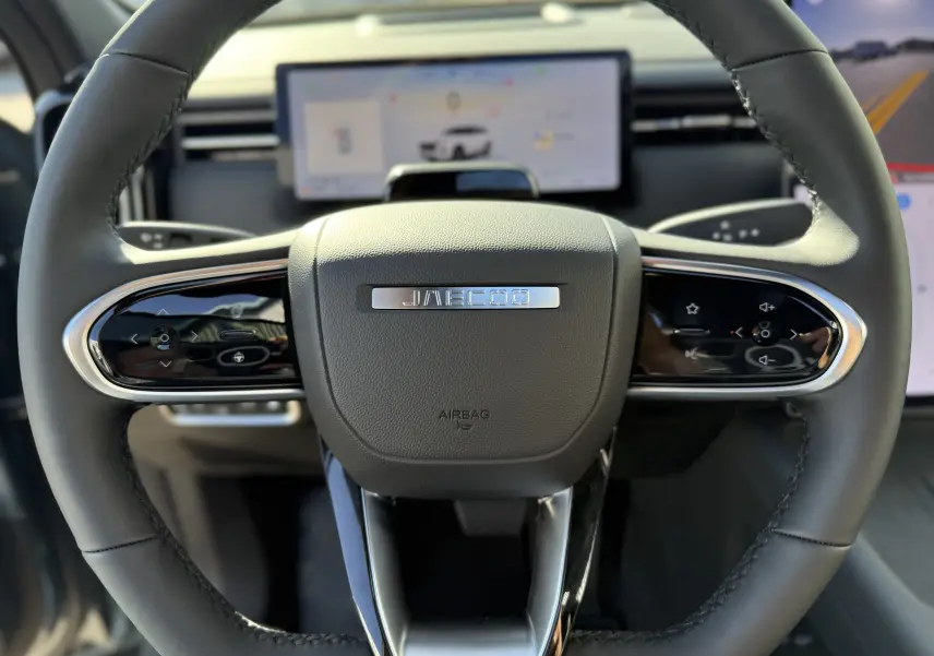 Vue rapprochée du volant noir du JAECOO J7 PHEV 2026 avec commandes tactiles et écran numérique flou en arrière-plan.