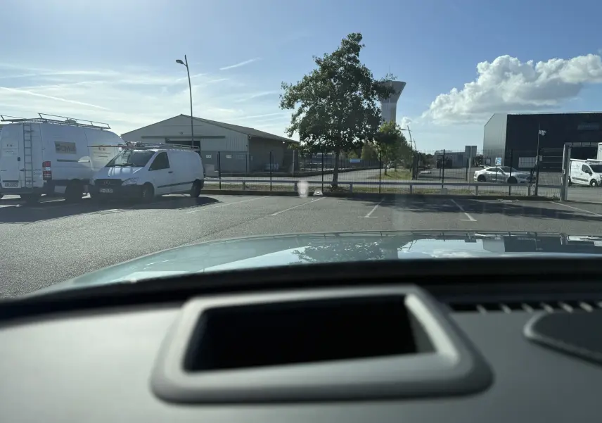 Vue depuis l'intérieur du JAECOO J7 blanc khaki, montrant le capot et un parking extérieur ensoleillé.