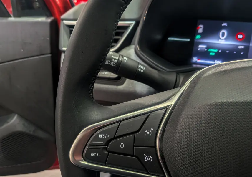 Gros plan sur le volant côté gauche de la Renault Clio V rouge Flamme, avec commandes de régulateur et tableau de bord numérique.