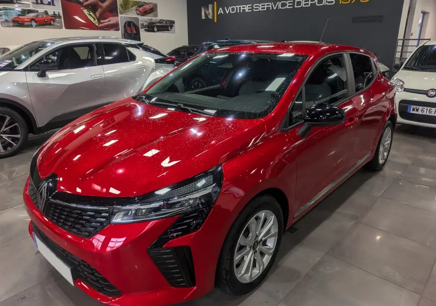 Renault Clio V rouge flamme vue 3/4 avant droit en intérieur, mettant en valeur ses phares LED et jantes alliage.