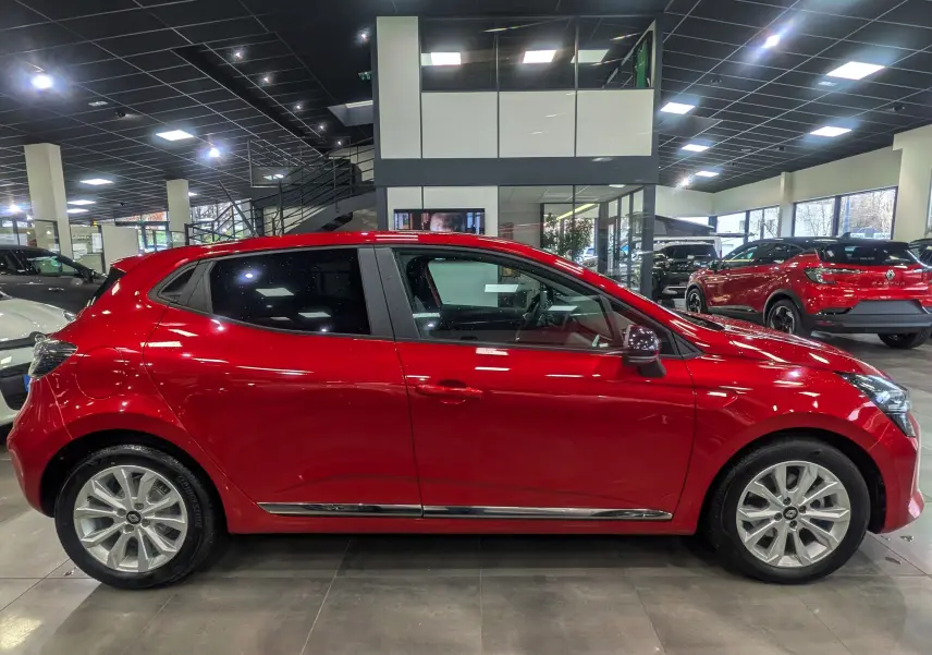 Profil latéral droit d'une Renault Clio V Rouge Flamme dans un showroom, soulignant ses jantes alliage et ses lignes fluides.