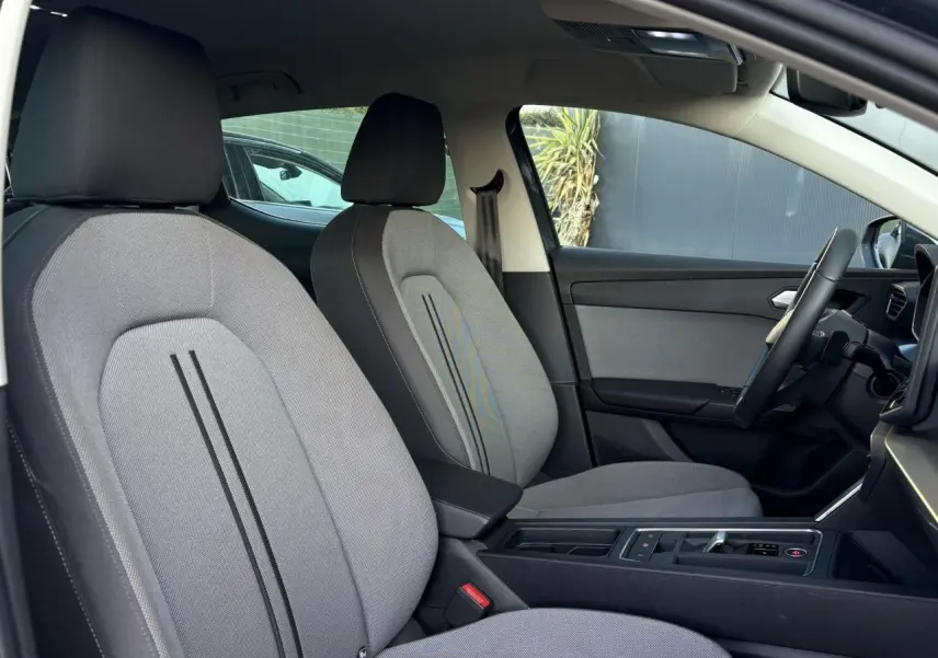 Vue intérieure côté conducteur du SEAT LEON 1.5 eTSI 115 DSG Style 2025, sièges tissu gris et accoudoir central noir.