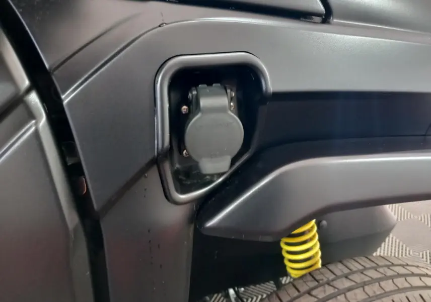 Détail de la prise de recharge électrique côté gauche d'une Aixam Mega E-Scouty Evo bleu, avec ressort de suspension jaune visible.