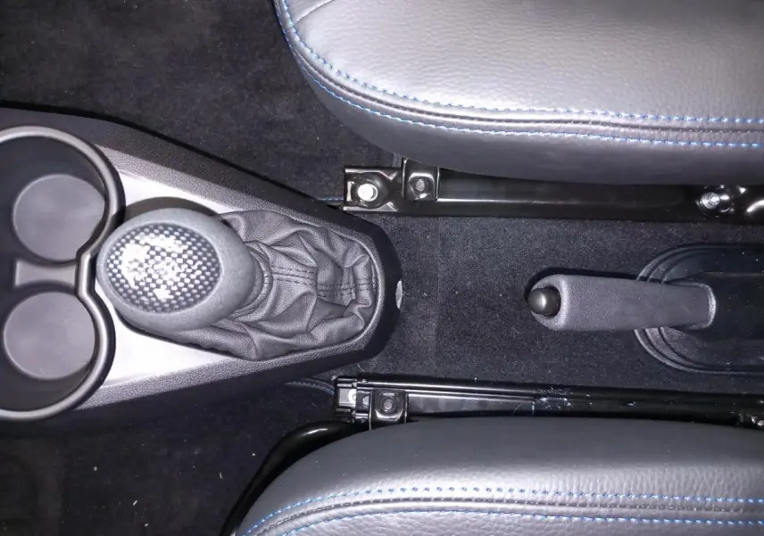 Vue intérieure centrée sur le levier de vitesse et frein à main entre deux sièges gris avec surpiqûres bleues.