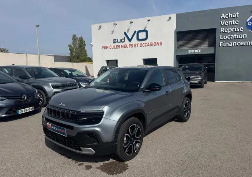 Jeep Avenger gris foncé en 3/4 avant droit, avec calandre noire distinctive et jantes modernes, stationné devant un concessionnaire.