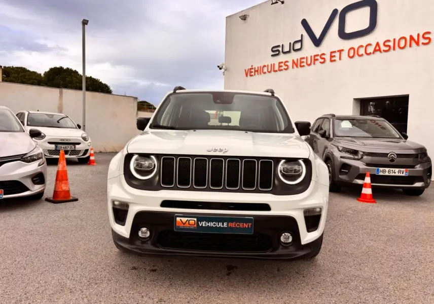 Vue frontale d'un Jeep Renegade blanc 2024 avec calandre noire et phares ronds allumés, stationné devant un concessionnaire.