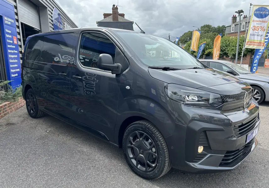 Vue 3/4 avant d'un Citroën Jumpy gris titane avec jantes noires, garé devant un garage en extérieur.