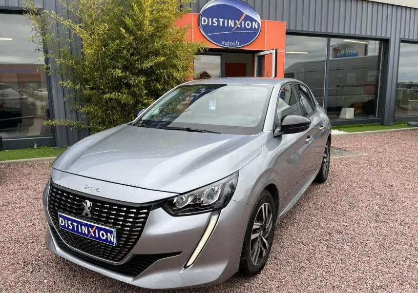 Peugeot 208 gris clair vue 3/4 avant, avec calandre noire et feux LED distinctifs, stationnée devant un bâtiment.