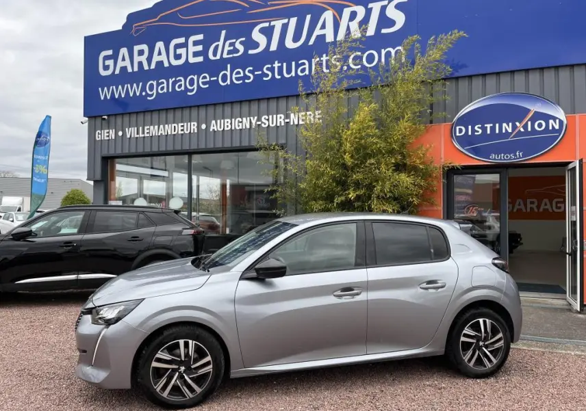 Peugeot 208 gris clair vue de profil côté gauche, stationnée devant un garage avec vitres arrière surteintées.
