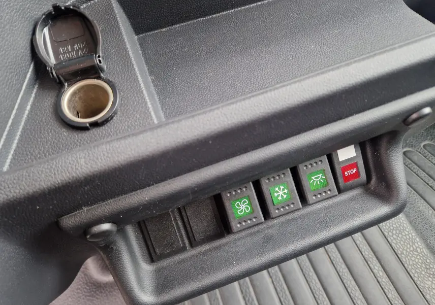 Gros plan sur les commandes vertes et rouge du tableau de bord noir du Renault Master blanc TPMR, avec prise allume-cigare ouverte.