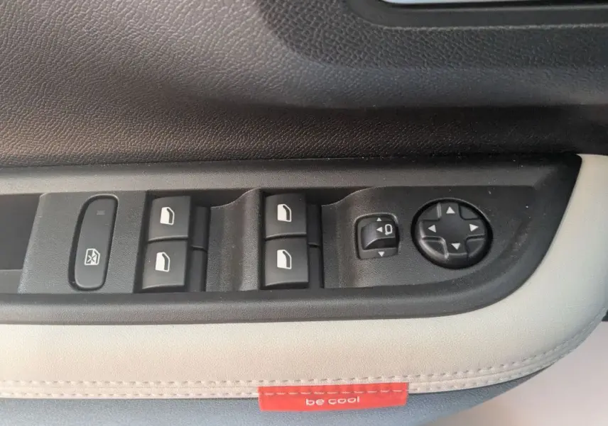 Gros plan sur les commandes de lève-vitres et réglage des rétroviseurs côté conducteur, sellerie claire avec étiquette rouge "be cool".