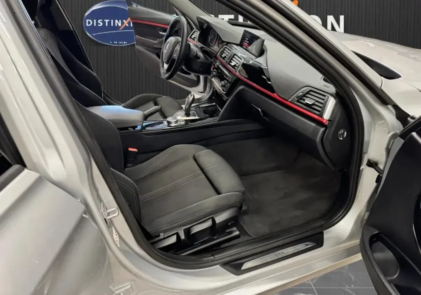 Vue intérieure côté conducteur d'une BMW Série 3 Touring gris aluminium, montrant sièges noirs et tableau de bord avec bande rouge.