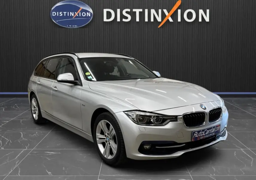 BMW Série 3 Touring F31 gris aluminium vue 3/4 avant droit, stationnée devant un mur noir avec logo Distinxion.