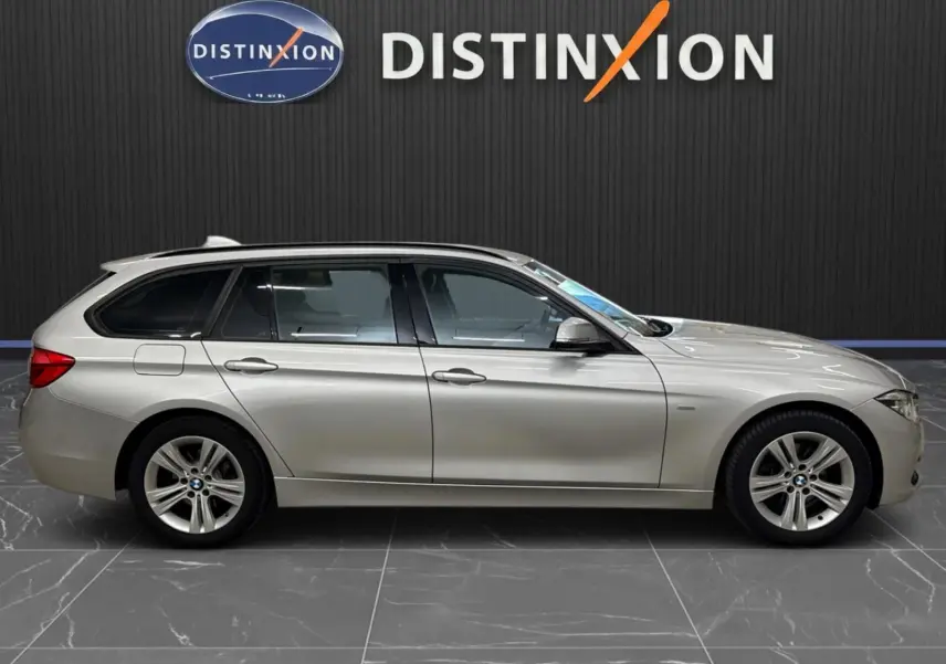 BMW Série 3 Touring F31 gris aluminium vue de profil côté gauche, stationnée devant un mur noir avec logo Distinxion.