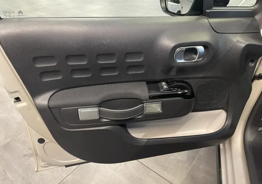 Vue intérieure côté gauche de la porte beige clair d'une Citroën C3 2024 avec accoudoir textile noir et poignée argentée.