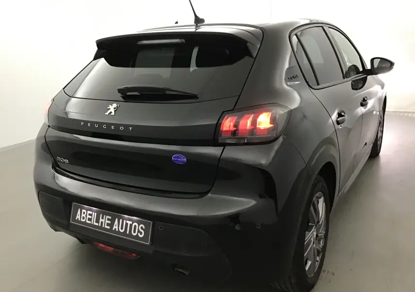 Vue 3/4 arrière droite d'une Peugeot 208 noire 2022 avec feux arrière allumés dans un garage blanc.