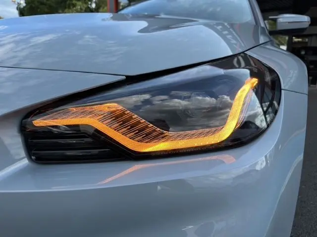 Gros plan sur le phare avant droit allumé d'une BMW M2 G87 gris Brooklyn Grau avec détails LED distinctifs.