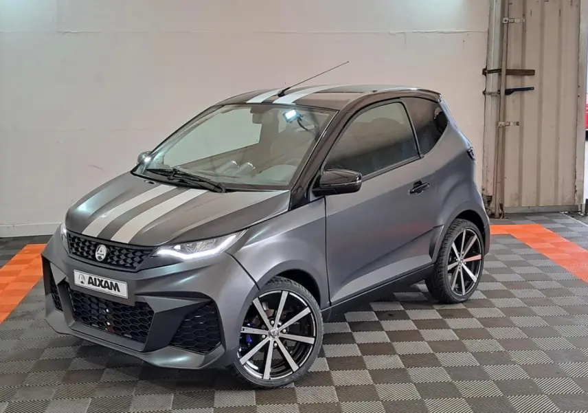 Vue 3/4 avant droite d'une Aixam Ambition gris clair avec bandes blanches, jantes alliage noires diamantées et clignotants à défilement.