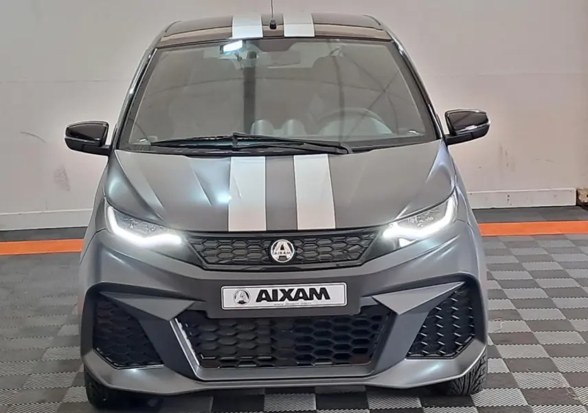 Vue avant d'une Aixam Ambition 2024 gris clair avec bandes blanches et phares LED allumés.