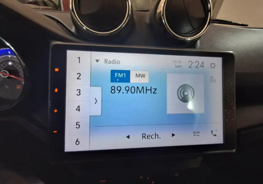 Écran tactile central de l’Aixam Ambition 2024 affichant la radio FM à 89,90 MHz, avec aérateurs ronds au-dessus.