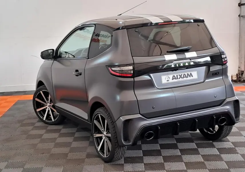 Vue 3/4 arrière droite d'une Aixam Ambition gris clair avec jantes alliage noires diamantées et bandes blanches sur le toit.