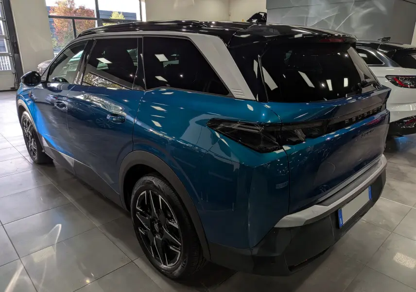 Vue 3/4 arrière droite du Peugeot 5008 Hybrid 145 GT bleu Obsession avec toit noir et jantes noires en intérieur showroom.
