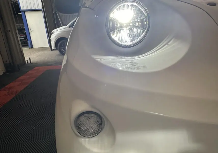 Gros plan sur le phare avant droit allumé de l'AIXAM MINAUTO E ACCESS blanc 2026, avec feu antibrouillard visible.