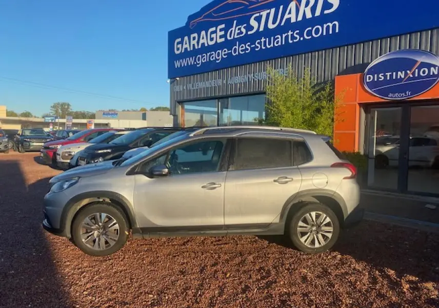 Peugeot 2008 gris Artense vue de profil côté gauche, stationnée devant un garage avec jantes aluminium 16 pouces.