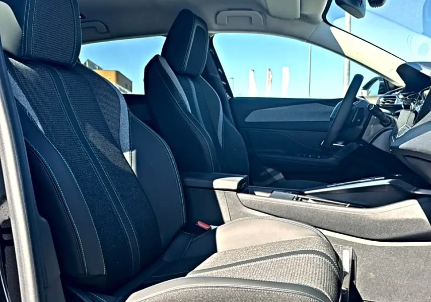 Vue intérieure côté gauche des sièges avant noirs avec accoudoir central et volant cuir de la Peugeot 408 Hybrid 145 allure 2025.