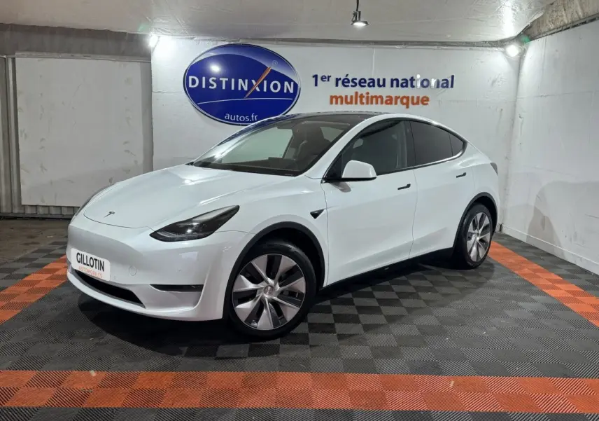 Tesla Model Y blanc en vue 3/4 avant droit, stationné sur sol à damier noir et orange en intérieur showroom.