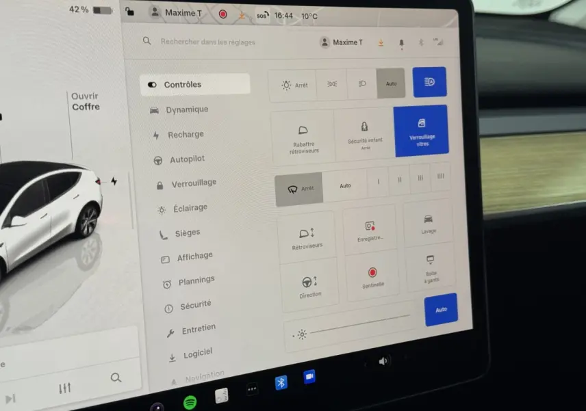Écran tactile central affichant les réglages du Tesla Model Y blanc, vue intérieure côté passager avec tableau de bord bois.