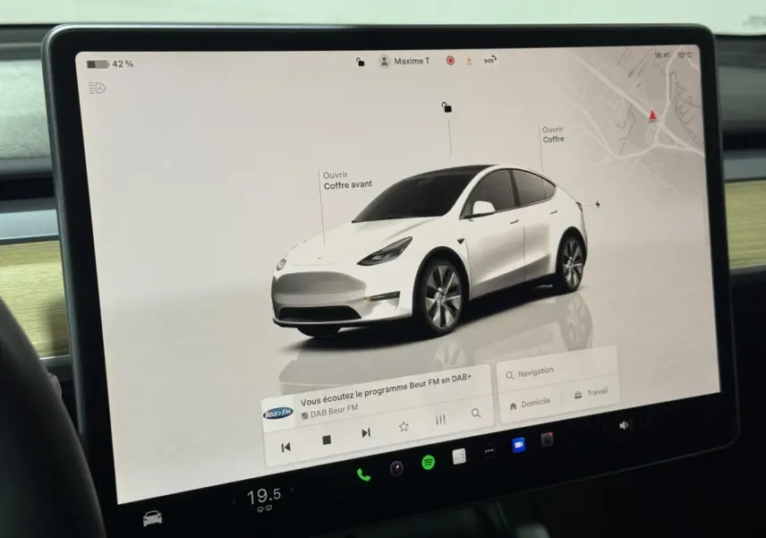 Vue 3/4 avant d’un Tesla Model Y blanc affiché sur l’écran tactile central avec options d’ouverture des coffres.