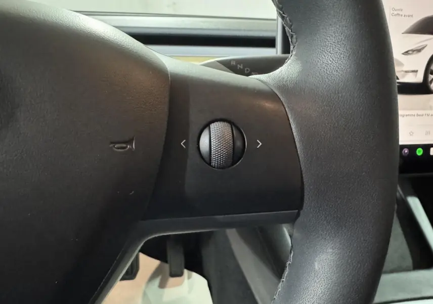 Gros plan sur le volant noir en cuir synthétique de la Tesla Model Y blanc, avec molette de contrôle tactile visible.