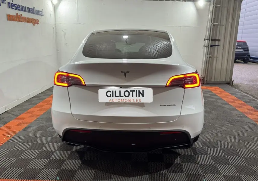 Vue arrière d'une Tesla Model Y blanc nacré avec feux arrière allumés dans un garage éclairé.