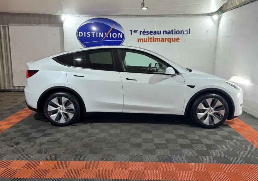 Profil côté gauche d'un Tesla Model Y blanc 2022 garé en intérieur sur sol damier gris et orange.