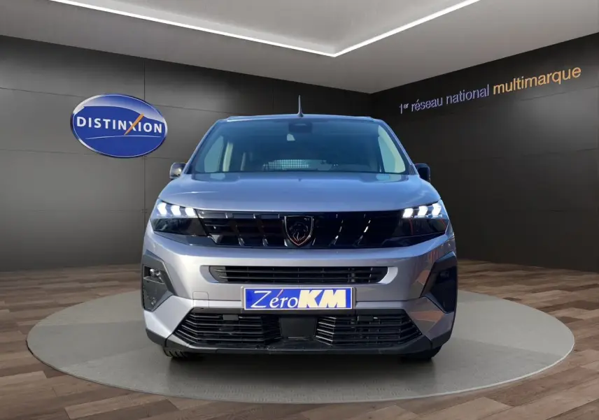 Vue frontale d'un Peugeot Rifter gris acier 2026 avec calandre noire et phares LED allumés en showroom.
