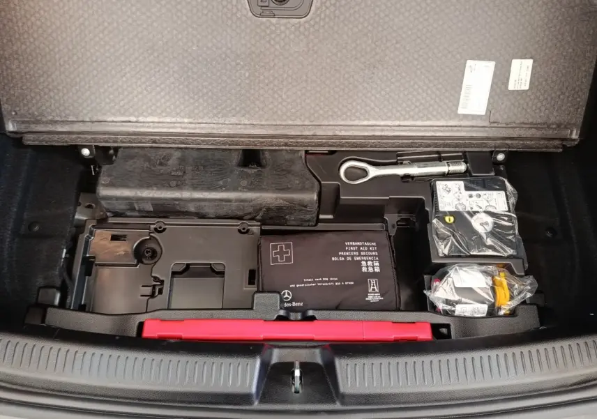 Vue plongeante sur le coffre ouvert d'une Mercedes Classe A noire, montrant la trousse de secours et outils rangés sous la tablette.