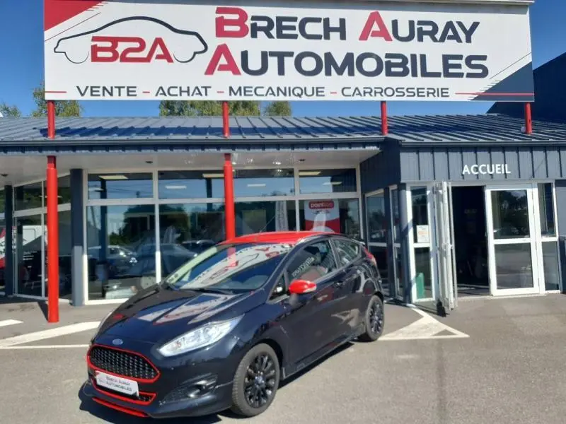 Ford Fiesta noire vue en 3/4 avant droit, avec rétroviseurs rouges et jantes noires, devant un garage Brech Auray Automobiles.