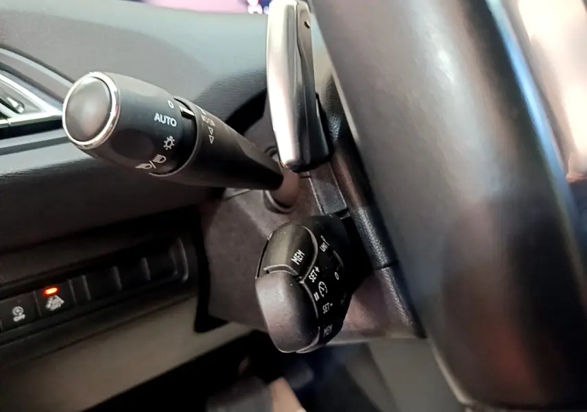 Gros plan sur les commandes au volant noir de la Peugeot 308 blanche, mettant en valeur les fonctions de régulateur et éclairage.