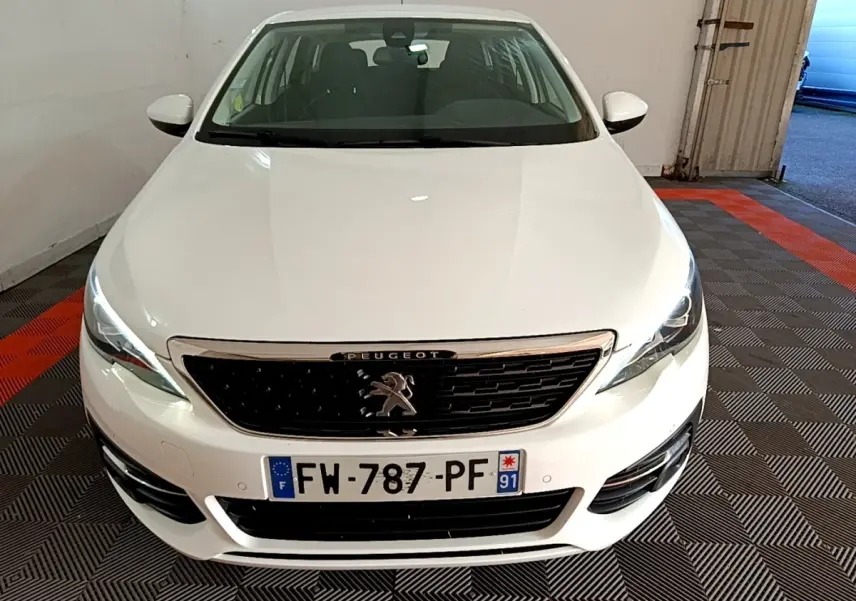 Vue de face d'une Peugeot 308 blanche avec calandre noire et phares LED allumés dans un garage.
